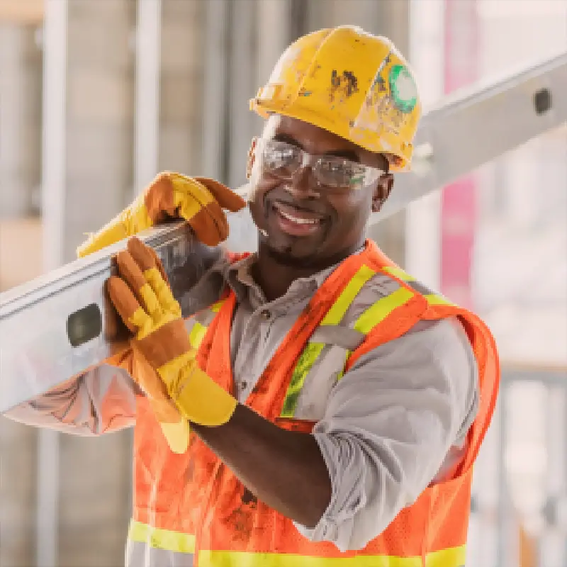Smiling construction worker in a yellow hard hat holding a spirit level at an active job site.